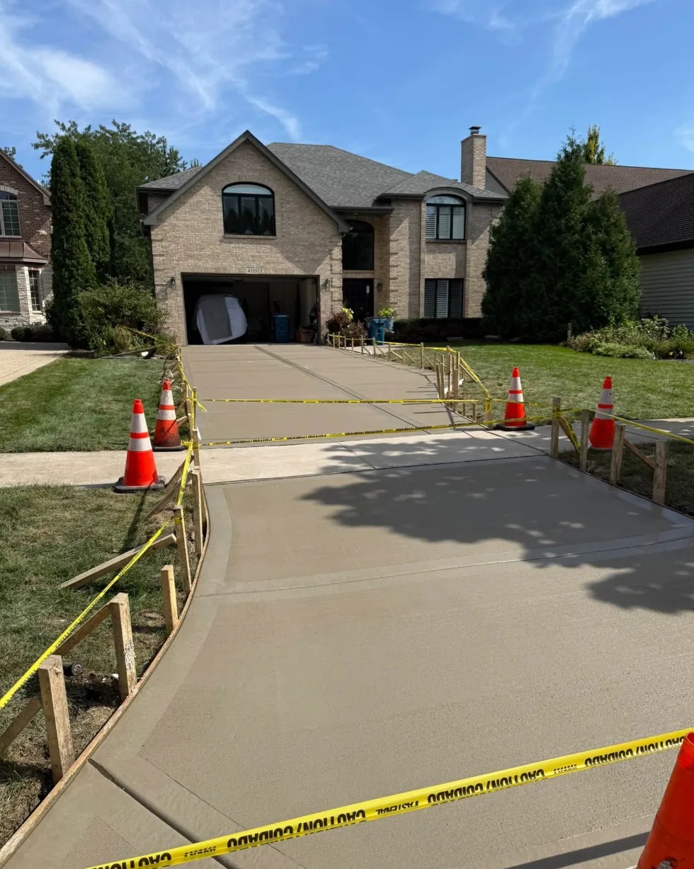 Freshly poured broom-finish concrete driveway running up to the attached garage of a two-story brick home, with safety cones and caution tape around the new pour