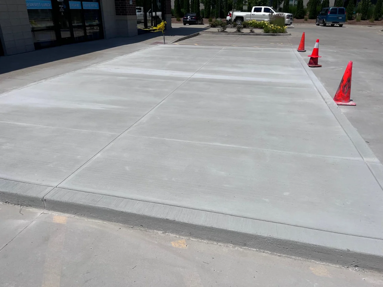 Large freshly poured concrete slab running along the front of a commercial storefront, cut into big square panels by control joints, with orange traffic cones and caution tape around the new pour and a parking lot behind it