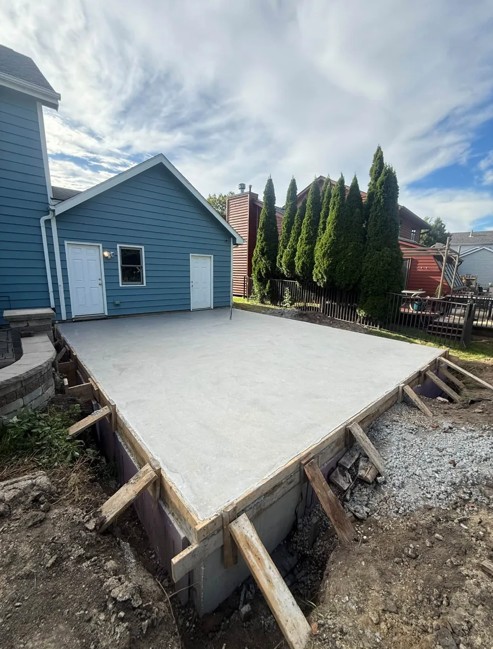 Newly poured elevated concrete slab beside a blue detached garage, with wood forms and the poured foundation wall still exposed along the edge