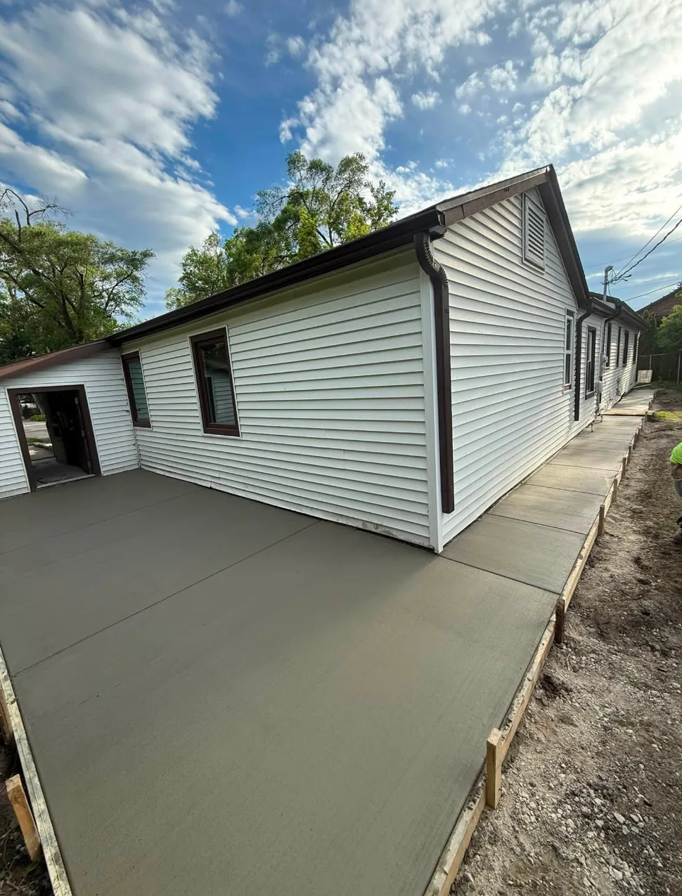 Large freshly poured concrete slab running the full length of a white-sided home, finished flat with wood form boards and stakes still set along the open edge