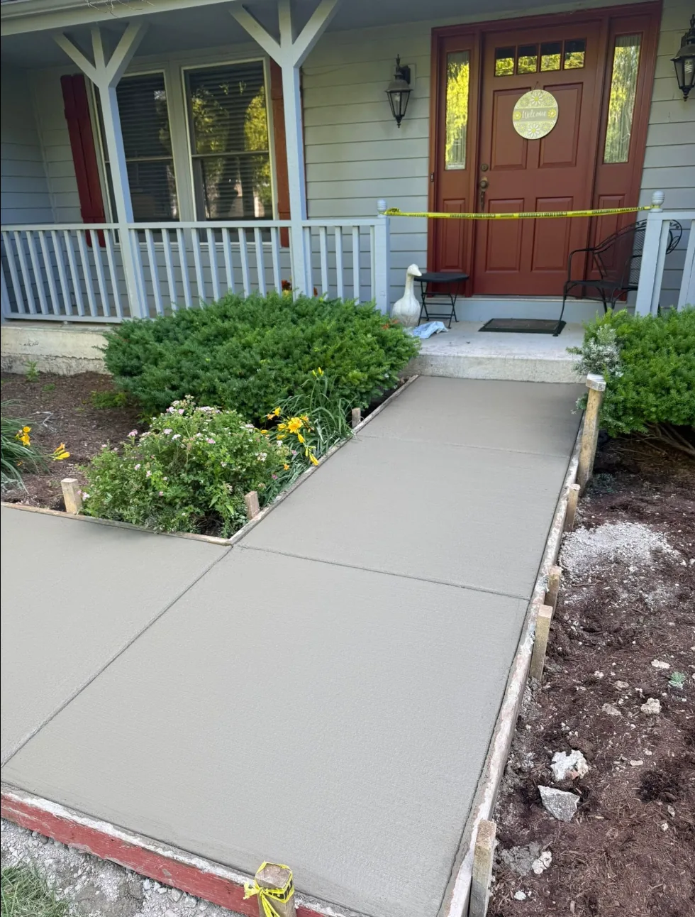 New broom-finish concrete walkway leading to the covered front entry of a gray-sided home, with crisp control joints and the wood edge forms still in place