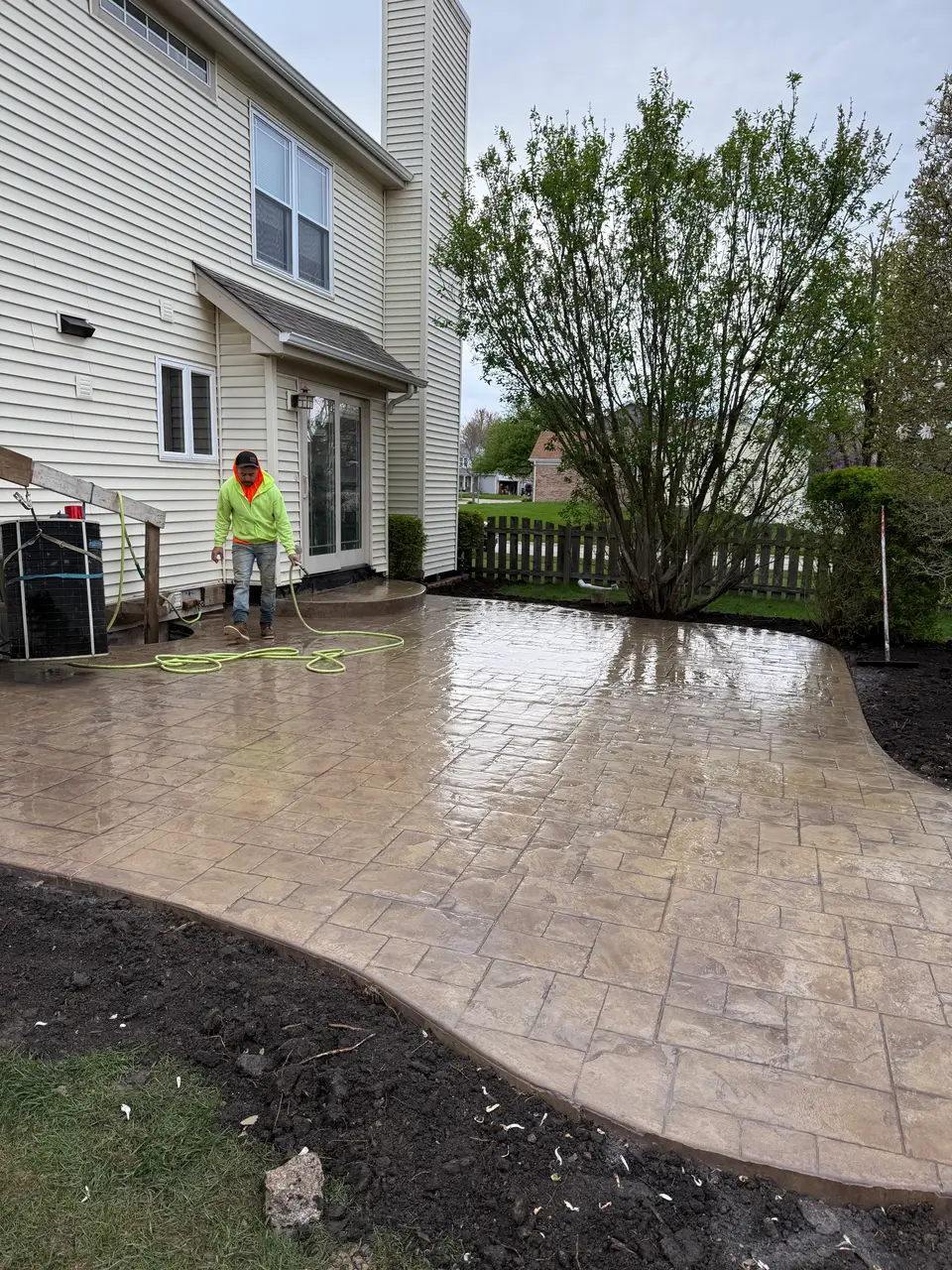 Freshly finished tan stamped concrete patio in a back yard, pressed in a random stone pattern with a curved border edge, being rinsed down with a hose by a worker in a hi-vis jacket beside a cream-sided two-story home